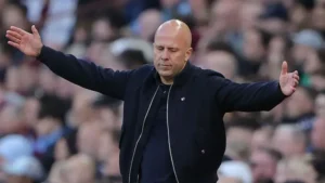 Bald man in a black jacket stands with arms spread wide on a sports field, eyes closed amid a cheering crowd behind him.