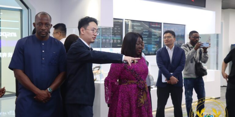 Presenter in a dark suit points to display screens in a bright showroom as a diverse group watches attentively.
