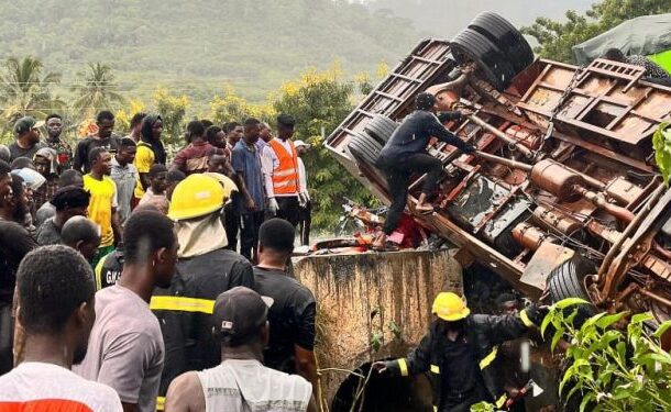 Crowd of onlookers and rescue workers surround a toppled truck as responders work to stabilize it near a roadside embankment.