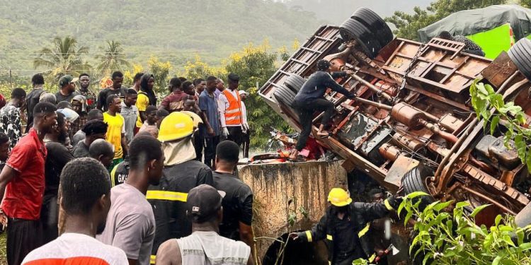 Crowd of onlookers and rescue workers surround a toppled truck as responders work to stabilize it near a roadside embankment.