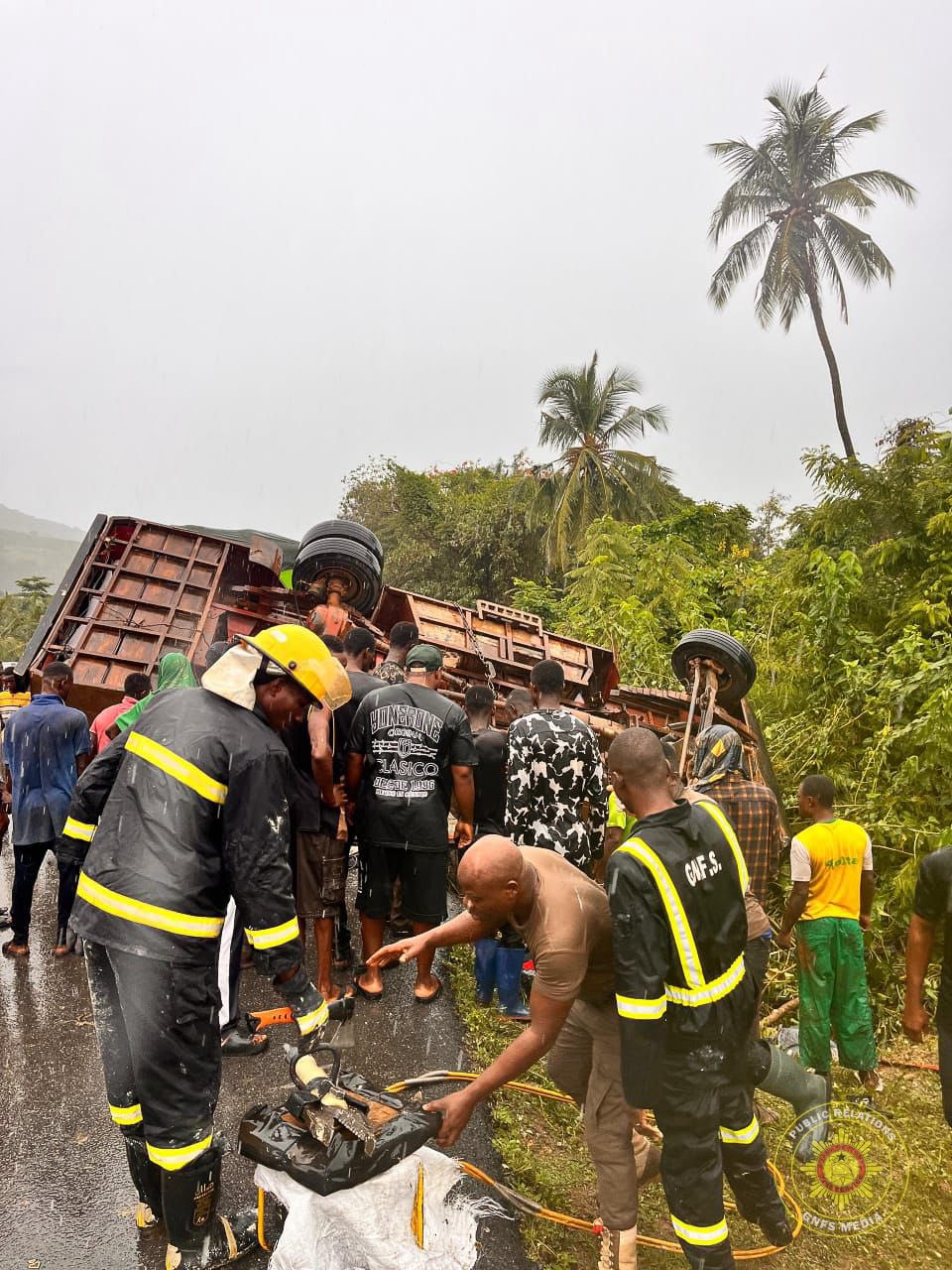 Rescue scene: firefighters and bystanders work around a overturned flatbed truck on a wet road during rain, with a fallen motorbike nearby and palm trees in the background.