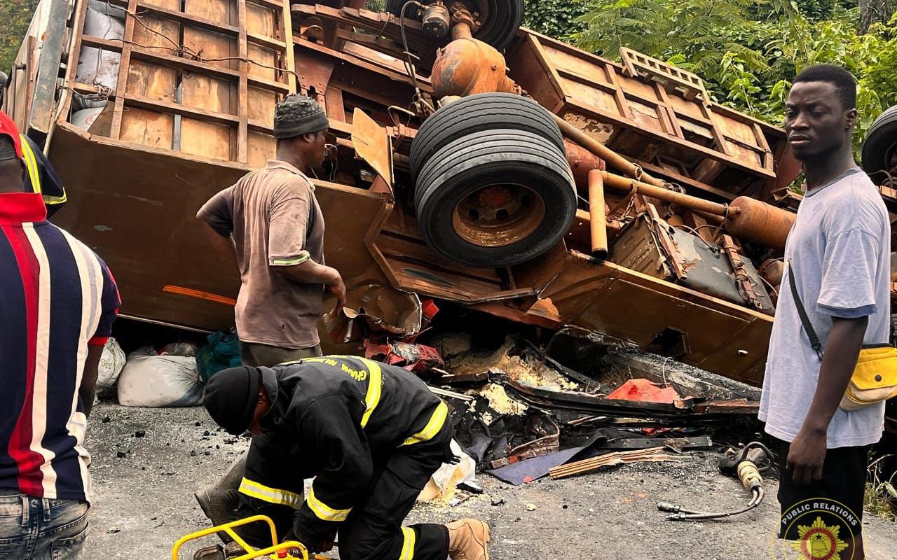 Emergency responders inspect a flipped, derailed truck with debris scattered on a road; a firefighter in gear kneels to assist.