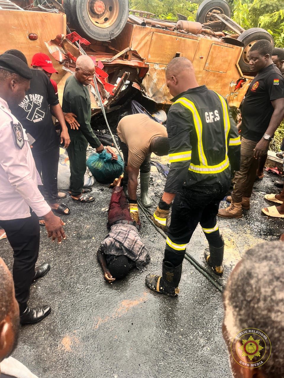 Rescue workers and bystanders crowd around a severely damaged overturned bus in a roadside crash, as one person lies on the ground being attended to and others pull debris away with a rope nearby.]}, {