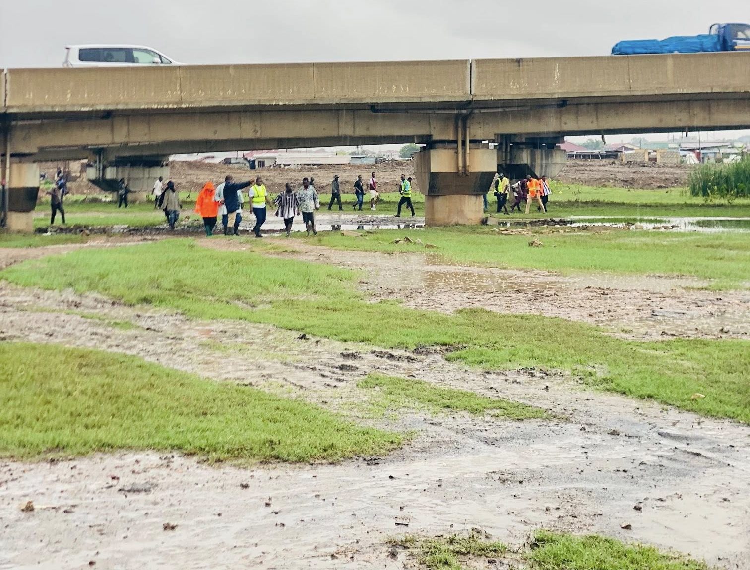 708345830_122173795970946649_28730242979836933_n-e1779958876886 | Ghanaguardian.com Group of people walking across a muddy field under a concrete bridge; several wear bright safety vests and jackets.