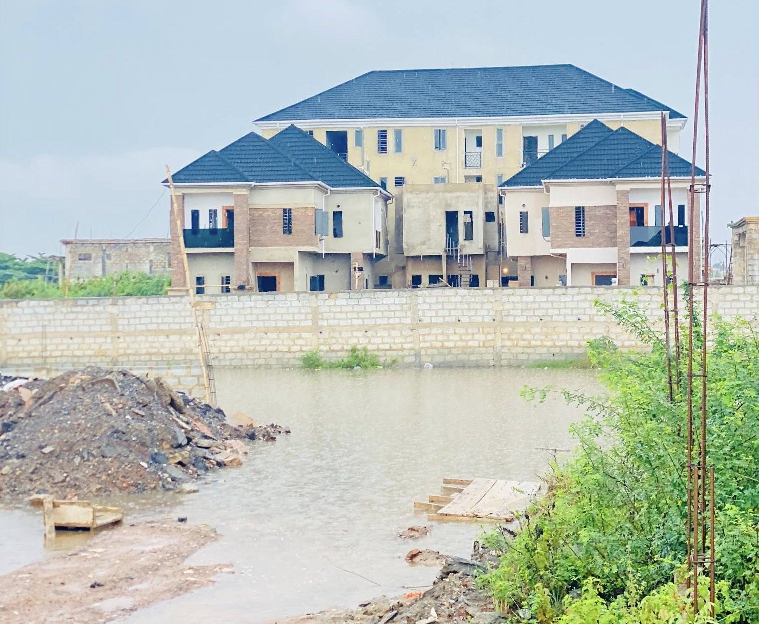 708363719_122173795988946649_7632310125969305891_n-e1779958932734 | Ghanaguardian.com Under-construction apartment complex behind a low brick wall, with a muddy foreground and rubble pile by a waterway in front of the site.