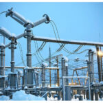 Snow-covered electrical substation with transformers, insulators, and overhead power lines against a blue sky.