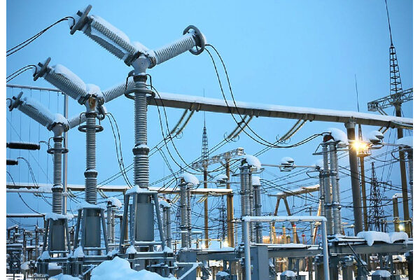 Snow-covered electrical substation with transformers, insulators, and overhead power lines against a blue sky.