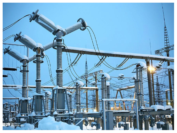 Snow-covered electrical substation with transformers, insulators, and overhead power lines against a blue sky.