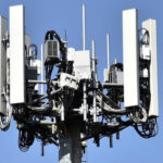 Cellular communication antenna array mounted on a pole against a blue sky, with multiple panel antennas and cables.