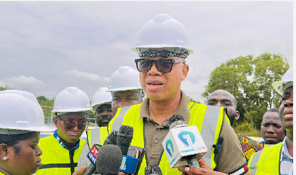 Man in a white hard hat and neon safety vest speaks to reporters, microphones held up, with a small crowd of workers behind him on a site.