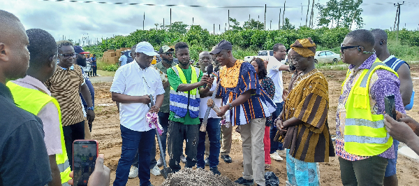 Group of people in safety vests and traditional attire gathered around a dirt mound at a construction site, likely a groundbreaking ceremony.