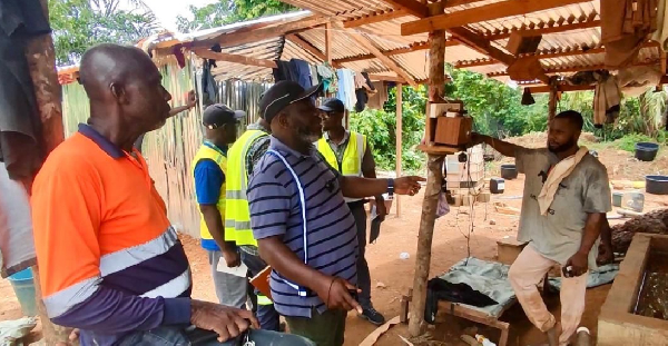 Group of men in a rustic workshop; one man in a striped shirt demonstrates or explains something to others, some wearing safety vests, under a wooden shelter with tools and hammocks nearby.