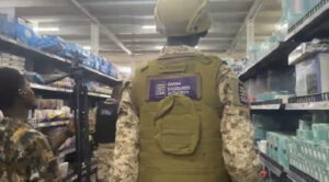 Back view of a uniformed officer in camouflage with a Ghana Standards Authority patch on the vest, walking down a supermarket aisle lined with shelves.