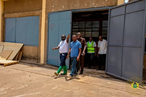 Group of men exit a warehouse through large blue doors, walking toward the camera; one in a blue polo leads while others follow.