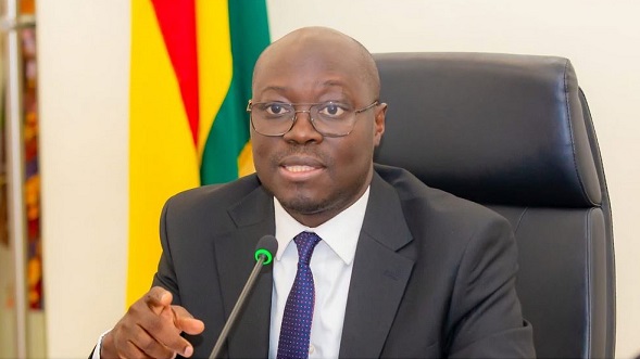 Professional man in a dark suit and tie speaks into a microphone at a desk, with colorful flags in the background.