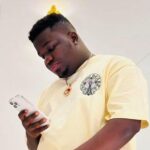 Young man in a pale yellow T-shirt looking at his smartphone indoors with a bright background.