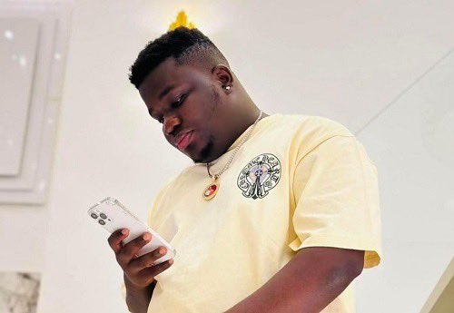 Young man in a pale yellow T-shirt looking at his smartphone indoors with a bright background.