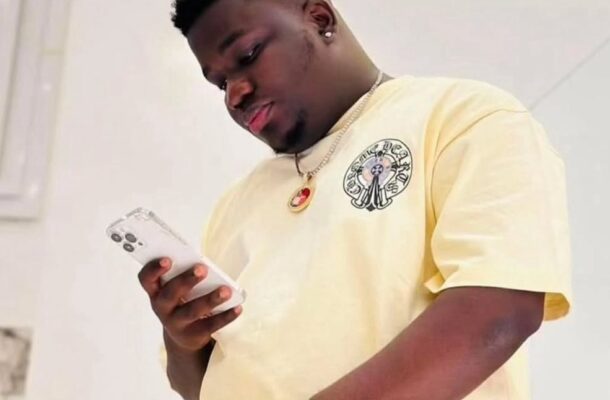 Young man in a pale yellow T-shirt with logo, looking at a white smartphone, wearing a silver watch and heart pendant necklace indoors.