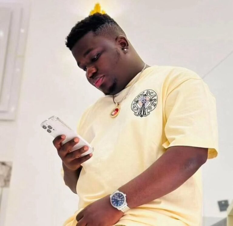 Young man in a pale yellow T-shirt with logo, looking at a white smartphone, wearing a silver watch and heart pendant necklace indoors.