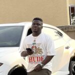 Young man standing beside a white SUV, holding a smartphone and looking at the camera, wearing a graphic t-shirt and cargo pants.