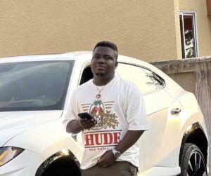 Young man standing beside a white SUV, holding a smartphone and looking at the camera, wearing a graphic t-shirt and cargo pants.