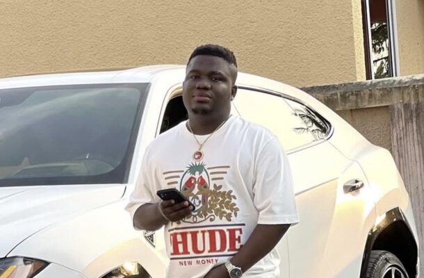 Young man standing beside a white SUV, holding a smartphone and looking at the camera, wearing a graphic t-shirt and cargo pants.