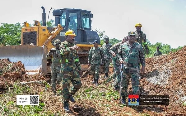 Ghana Armed Forces soldiers in camouflage with yellow helmets walk across a muddy construction site, a large bulldozer in the background.