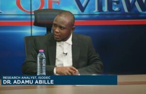 Dr. Adamu Abille, research analyst for ISODEC, sits at a TV news desk during an interview, with a water bottle on the table.