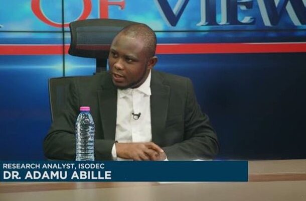 Dr. Adamu Abille, research analyst for ISODEC, sits at a TV news desk during an interview, with a water bottle on the table.