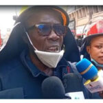 Firefighter wearing a helmet and sunglasses speaks to reporters on a street, microphones held up to him by journalists.