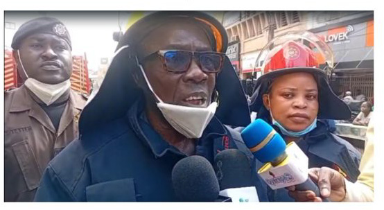 Firefighter wearing a helmet and sunglasses speaks to reporters on a street, microphones held up to him by journalists.