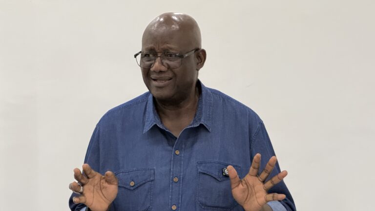 Man with glasses and a blue denim shirt gesturing with his hands while speaking, in mid-conversation.