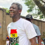 Joyful man in a white T-shirt with a red-yellow-green flag design and the word THE RISE, outdoors beside a wall, another man in a cap behind him.