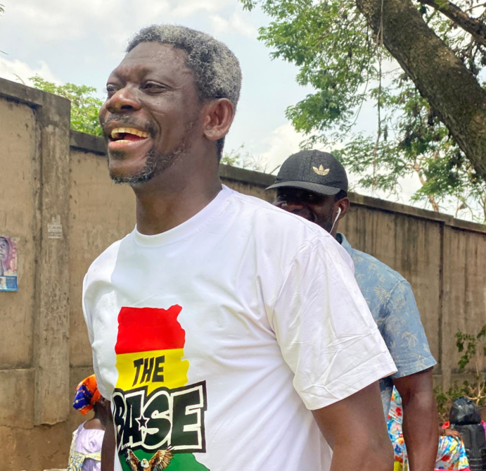 Joyful man in a white T-shirt with a red-yellow-green flag design and the word THE RISE, outdoors beside a wall, another man in a cap behind him.