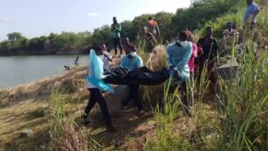 Group of responders carrying a dark body bag along a grassy riverbank, near the water's edge, with onlookers watching nearby.