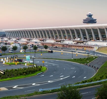Modern airport terminal with a long curved concourse, a circular road in the foreground, and a control tower in the distance at sunset.
