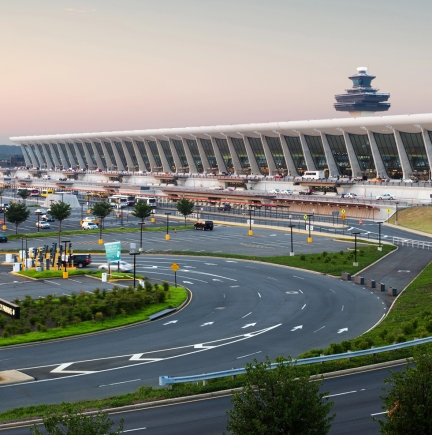 Modern airport terminal with a long curved concourse, a circular road in the foreground, and a control tower in the distance at sunset.