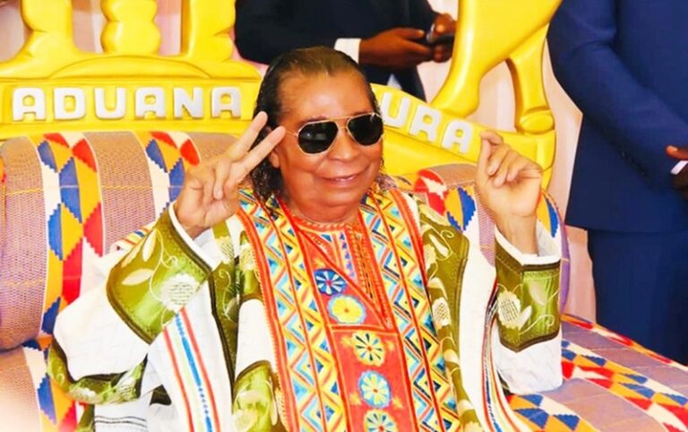 Older man in a bright, patterned robe and sunglasses sits on a colorful throne, flashing peace signs with both hands at a festive event.