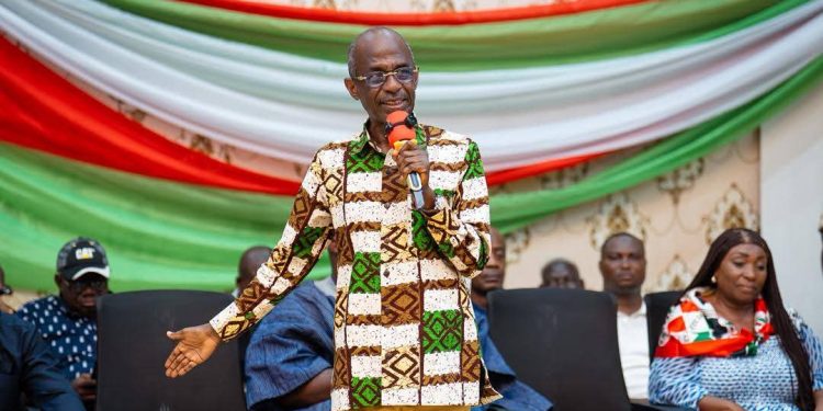 Man in a patterned green-brown shirt speaks into a microphone on a stage with red, white, and green drapes behind him.