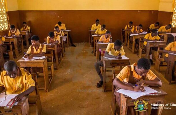 Students in yellow uniforms sit at wooden desks in a Ghanaian classroom, writing during an exam or assignment.