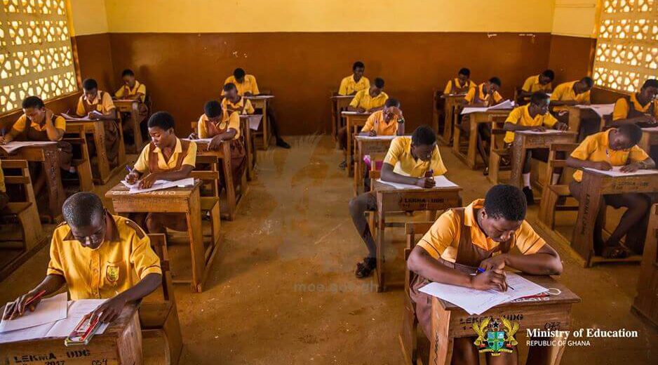Students in yellow uniforms sit at wooden desks in a Ghanaian classroom, writing during an exam or assignment.