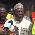 Man in a traditional cap and tunic speaks to reporters, microphones held up as others in high-visibility vests observe behind him.