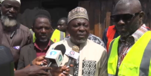 Man in a traditional cap and tunic speaks to reporters, microphones held up as others in high-visibility vests observe behind him.