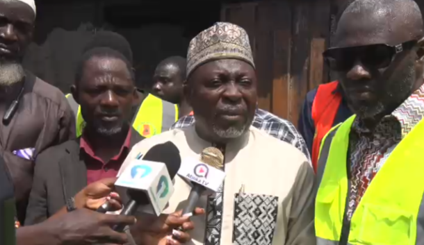 Man in a traditional cap and tunic speaks to reporters, microphones held up as others in high-visibility vests observe behind him.