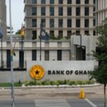 Front view of the Bank of Ghana building with a circular yellow emblem and the words 'BANK OF GHANA' on a curved wall, flags on flagpoles, and a pedestrian walking on the street in the foreground.
