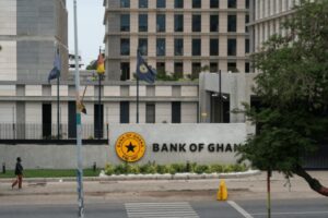 Front view of the Bank of Ghana building with a circular yellow emblem and the words 'BANK OF GHANA' on a curved wall, flags on flagpoles, and a pedestrian walking on the street in the foreground.