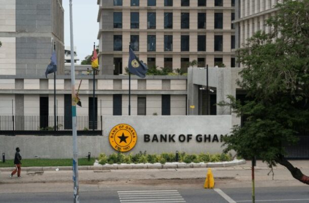 Front view of the Bank of Ghana building with a circular yellow emblem and the words 'BANK OF GHANA' on a curved wall, flags on flagpoles, and a pedestrian walking on the street in the foreground.