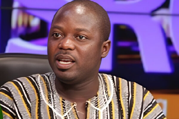 Close-up of a man in a striped shirt speaking on a TV talk show set with a purple backdrop.