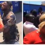 Two people arguing in a street; a woman in red blouse faces a man in camouflage as onlookers watch.