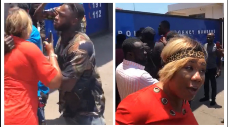 Two people arguing in a street; a woman in red blouse faces a man in camouflage as onlookers watch.